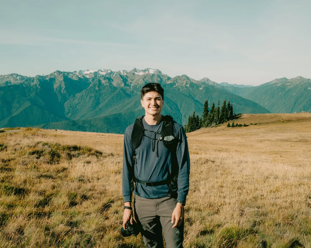 Omar Mussehl standing in a golden meadow below the Olympic mountains, camera in hand
